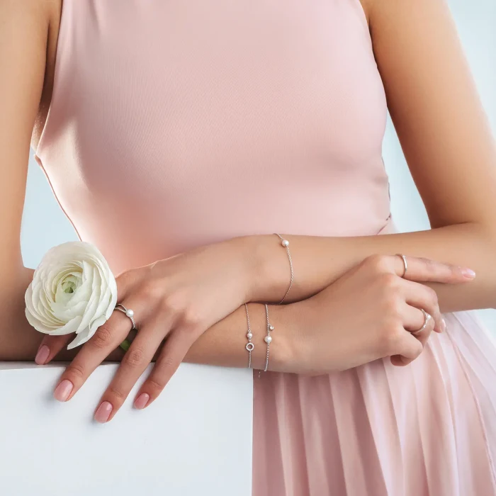GAURA SK22244B-2 A woman in a pink dress showcases elegant jewelry, holding a white flower, with delicate rings and bracelets accentuating her graceful hands.